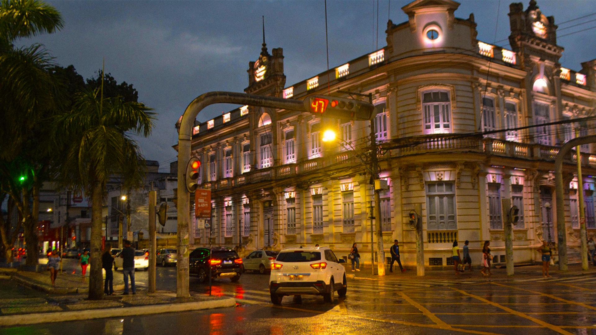 Centro histórico de Feira de Santana ao anoitecer, com o Paço Municipal iluminado e a movimentação urbana da avenida principal